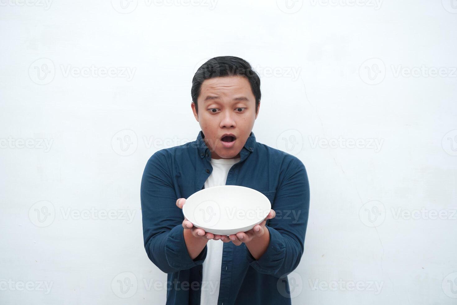 Portrait of happy Asian man wearing a blue shirt while holding an empty plate with happy and excited expression. Young man showing and pointing empty mockup plate. Isolated with a white background. photo