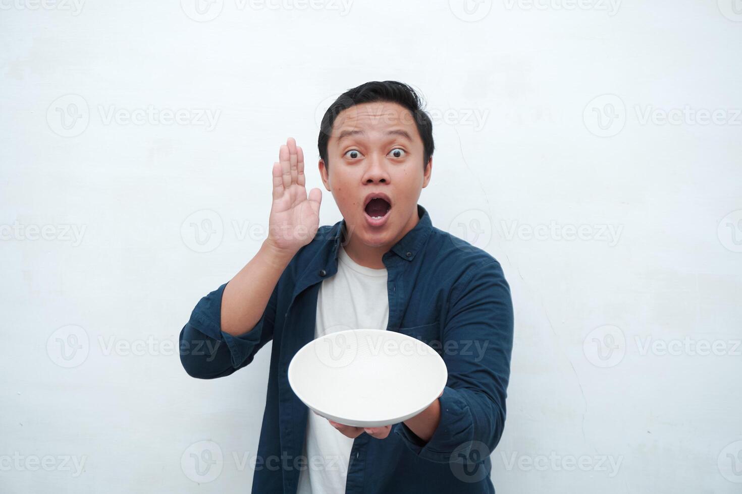 Portrait of happy Asian man wearing a blue shirt while holding an empty plate with happy and excited expression. Young man showing and pointing empty mockup plate. Isolated with a white background. photo