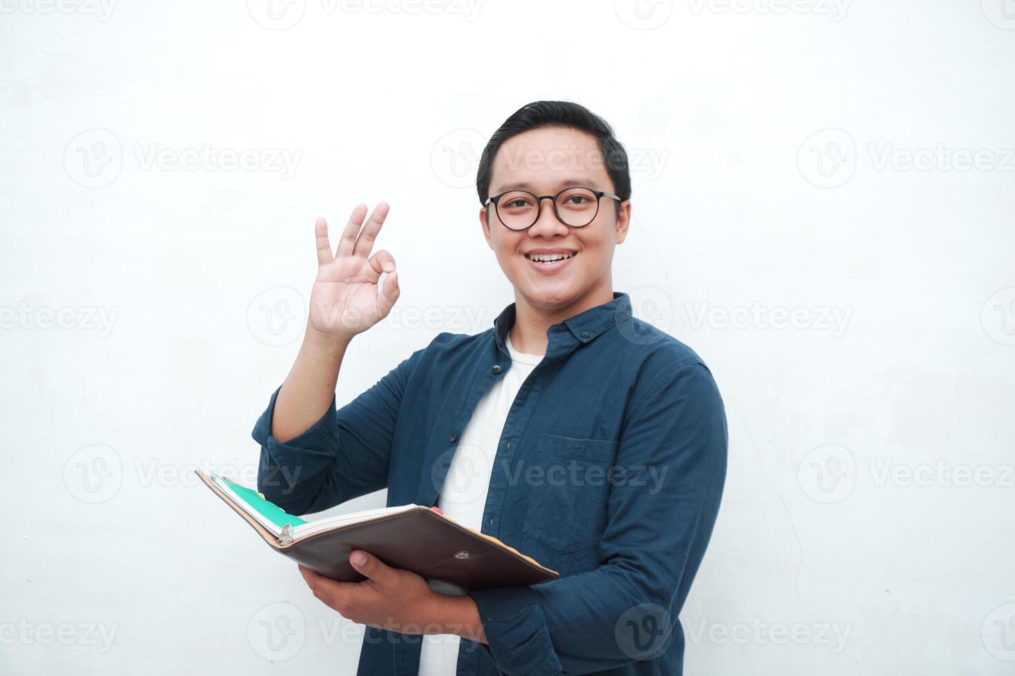 Young Asian man holding books smiling happy and positive. Asian student showing ok gesture and approval sign over white background photo