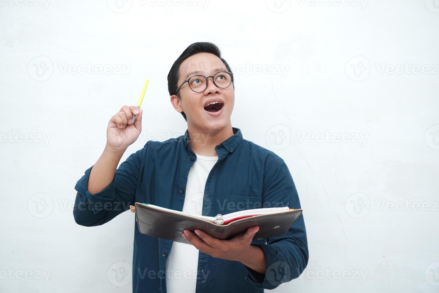 Happy young Asian man with glasses showing expression of gets genius idea. Asian college student showing happy and excited expression while holding book and pen photo