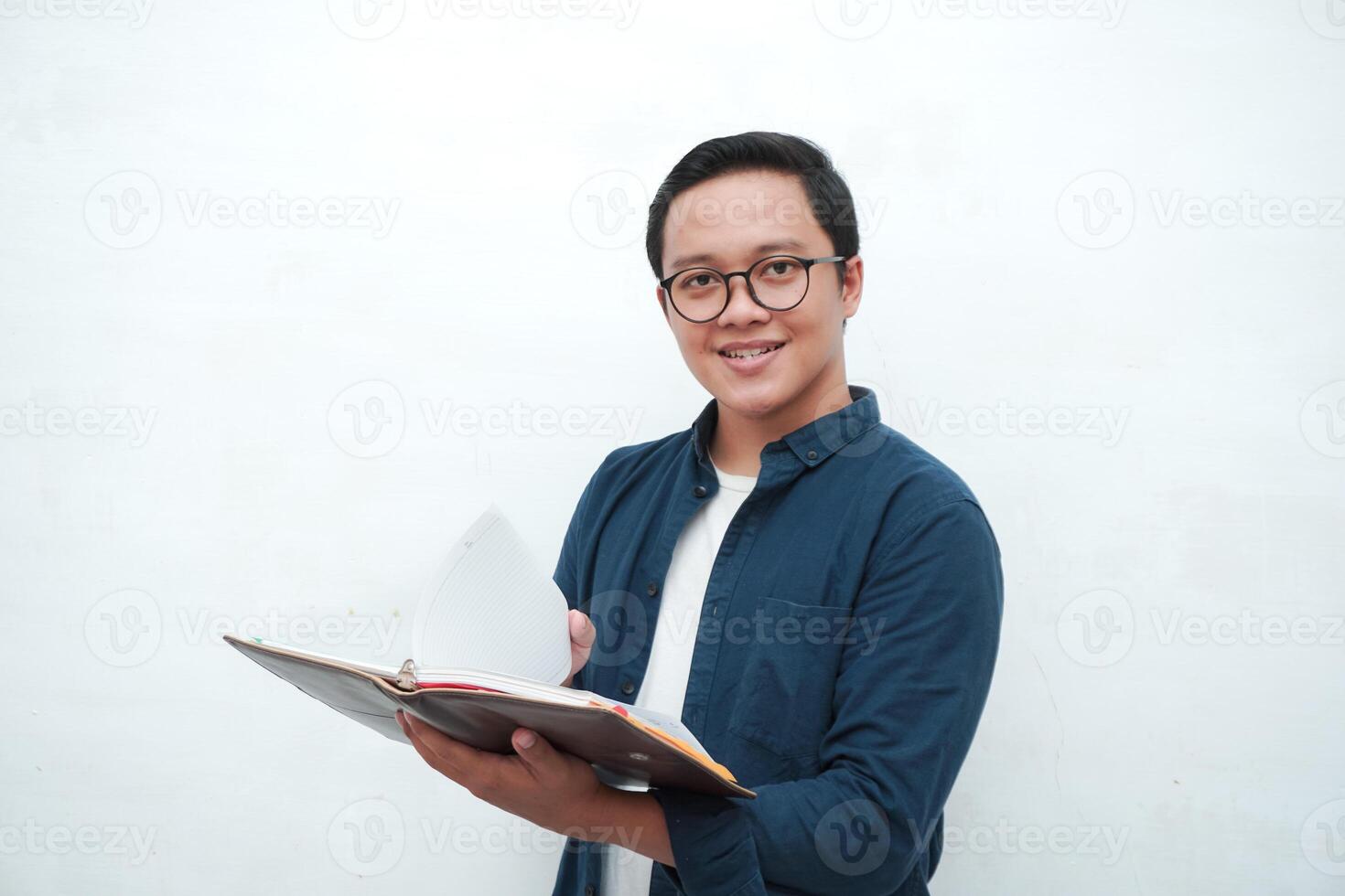 Young Asian man holding books smiling happy and positive while reading a book. Asian student flipping a book page over white background photo