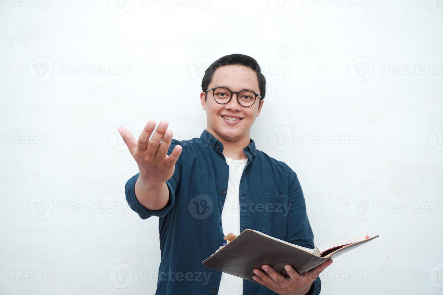 Young Asian man holding books smiling happy and positive while reading a book. Asian student pointing and showing welcoming gesture photo