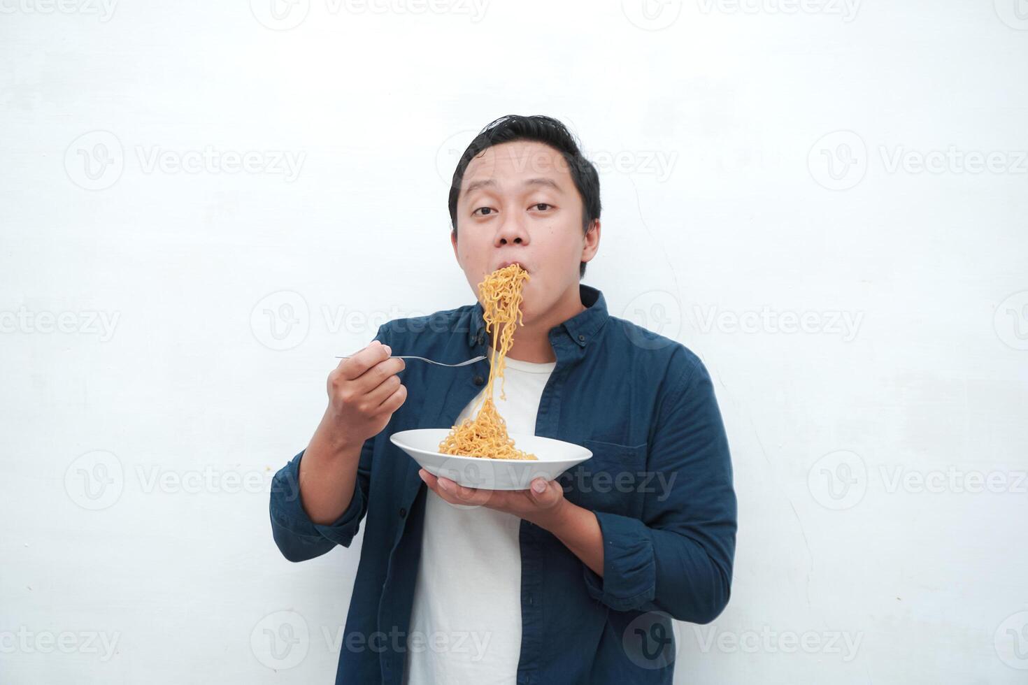 A portrait of happy Asian man wearing a blue shirt while holding a plate of fried noodles. Young man smiling and pointing to a plate of noodle. Isolated with a white background. photo