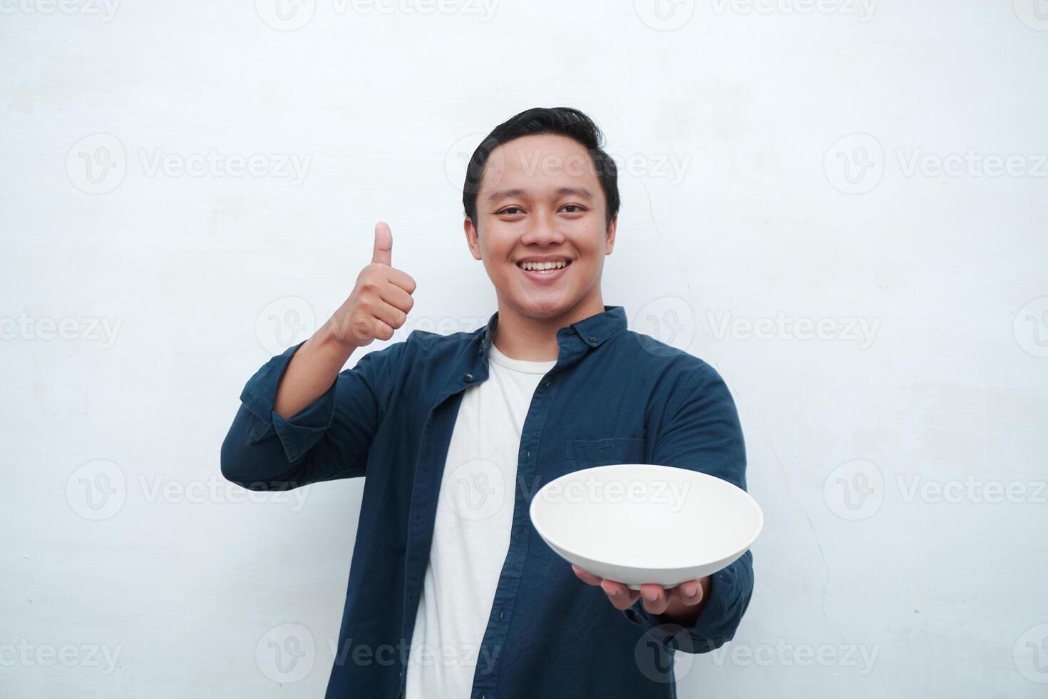 Portrait of happy Asian man wearing a blue shirt while holding an empty plate with happy and excited expression. Young man showing and pointing empty mockup plate. Isolated with a white background. photo
