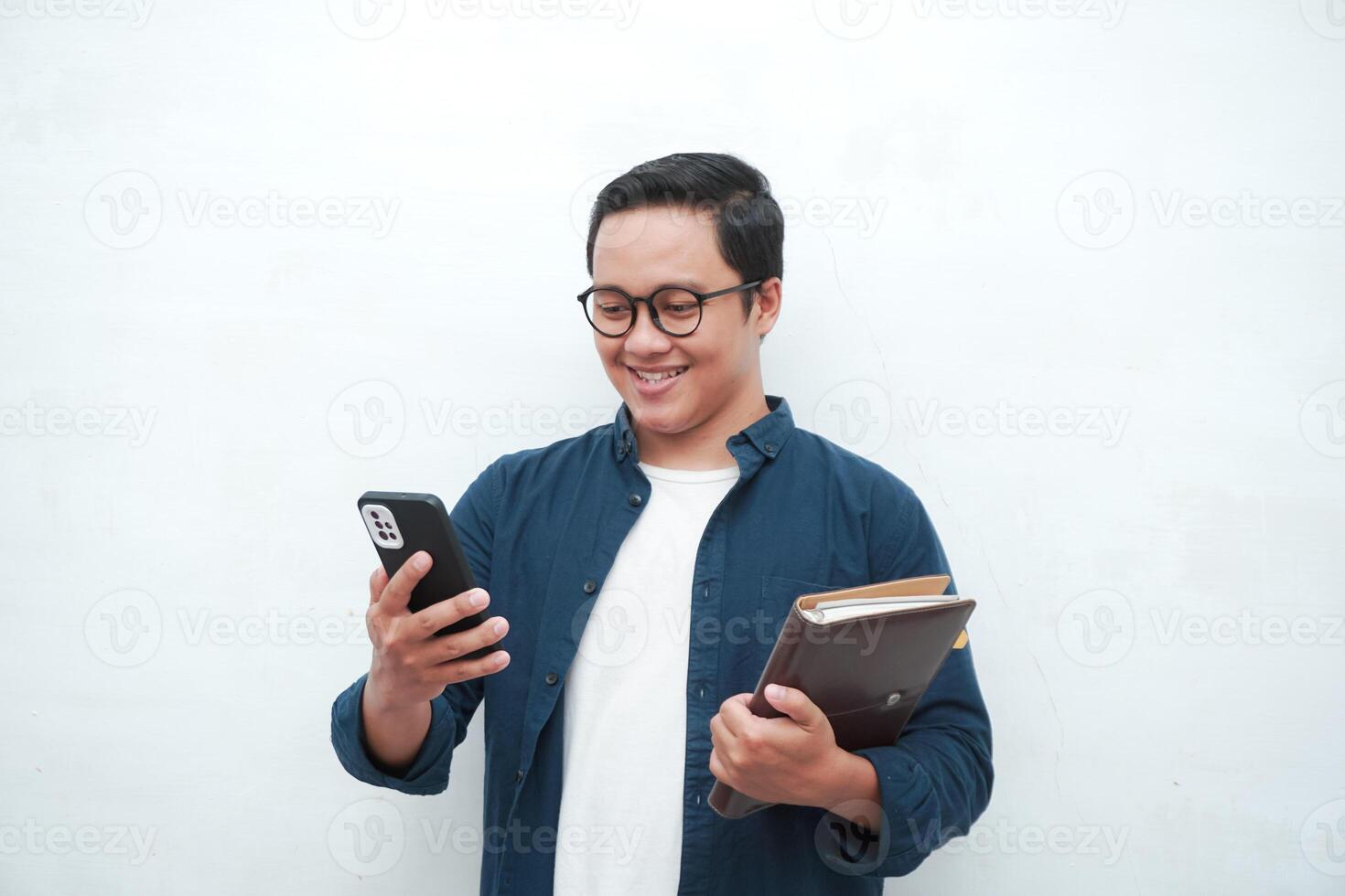 Asian college student holding book and blank smartphone screen with happy and excited expression. Young Asian student wearing blue shirt smiling and showing white mobile phone screen photo