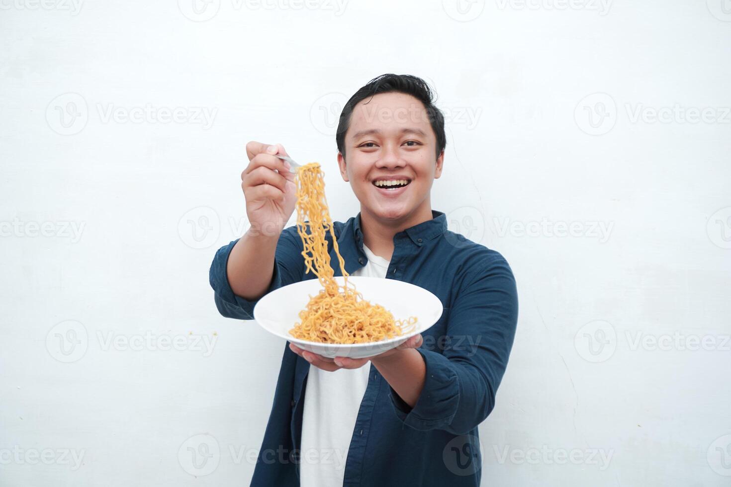 A portrait of happy Asian man wearing a blue shirt while holding a plate of fried noodles. Young man smiling and pointing to a plate of noodle. Isolated with a white background. photo