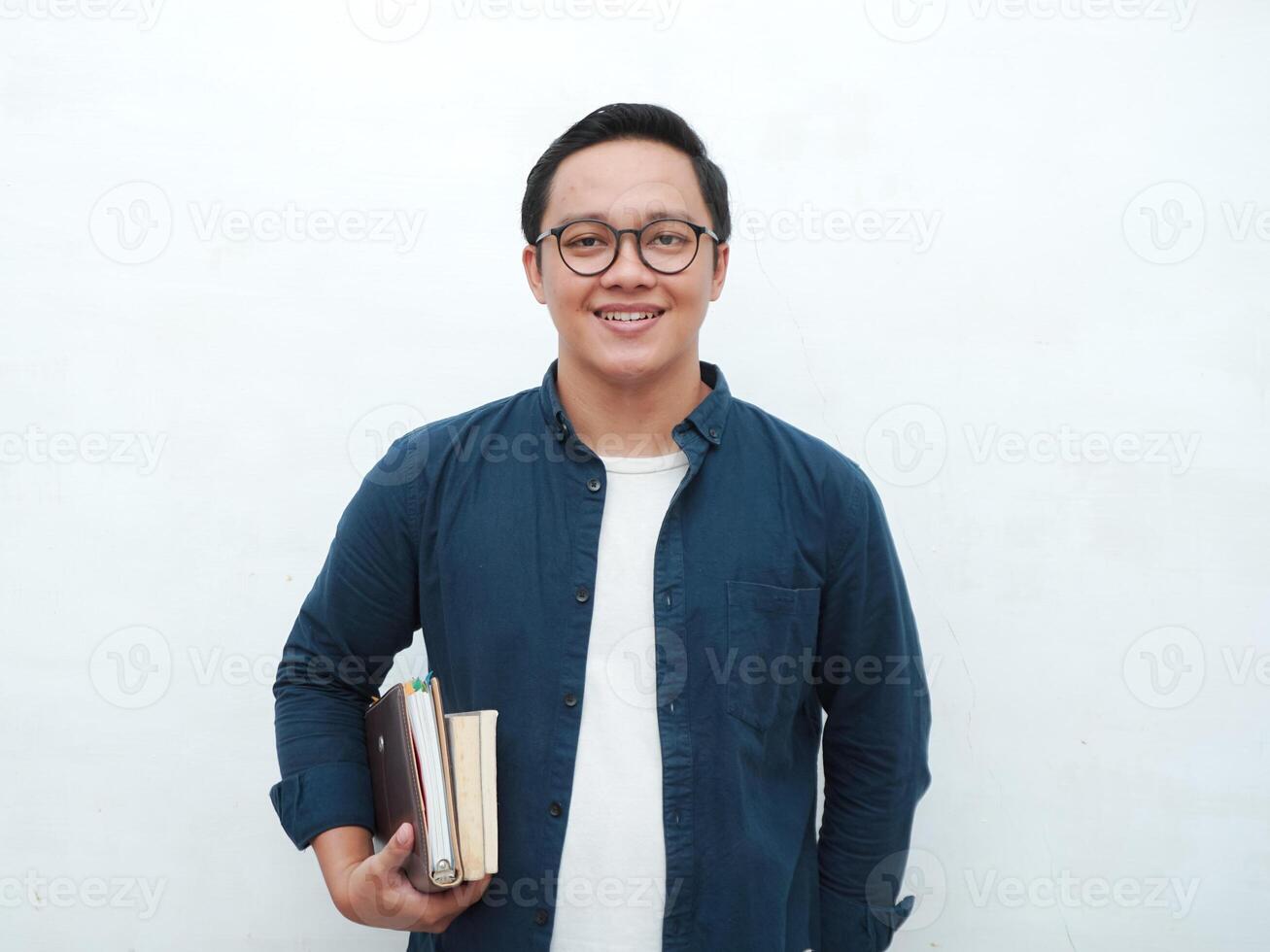 Young Asian man holding books smiling happy and positive while reading a book. Asian student flipping a book page over white background photo