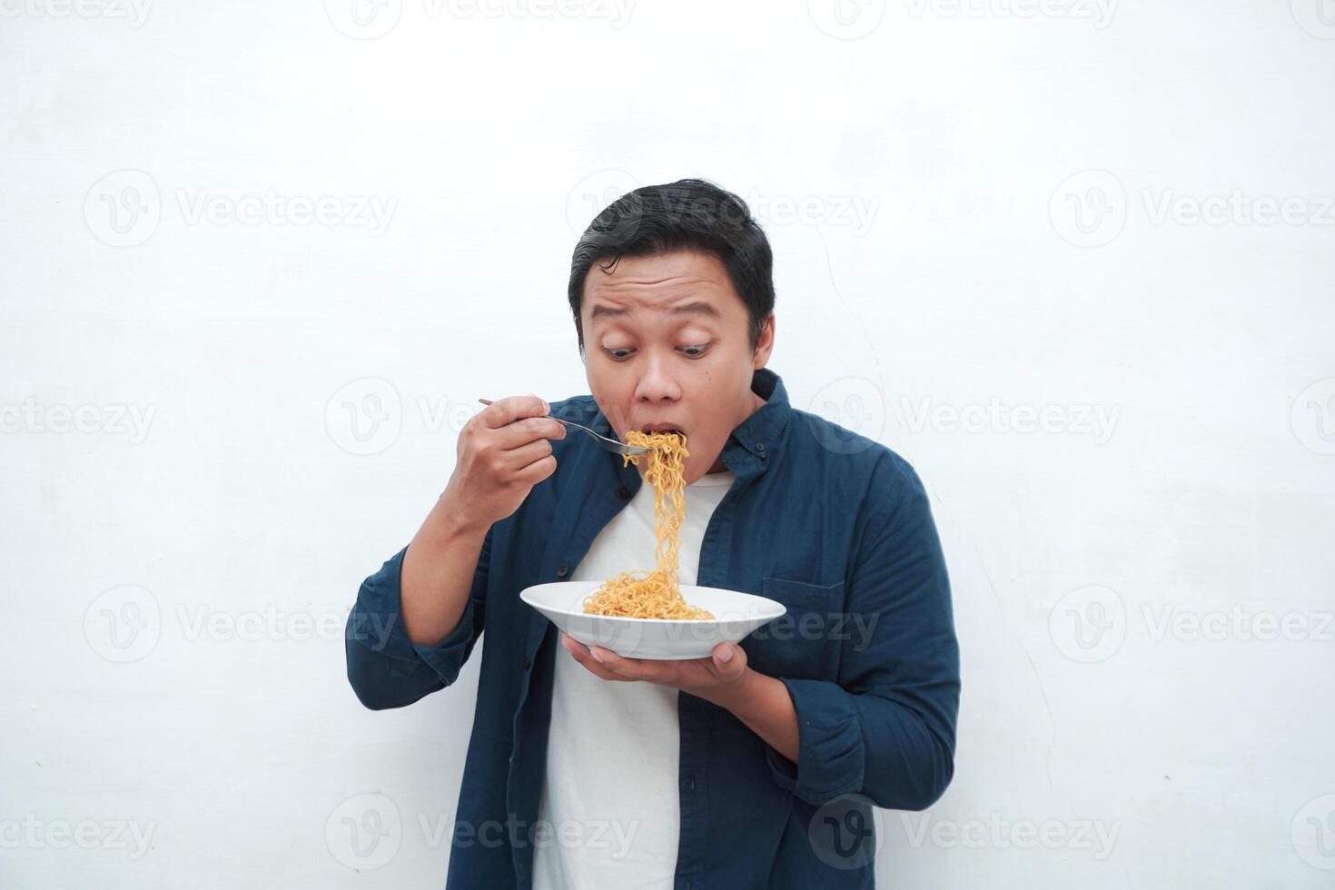 A portrait of happy Asian man wearing a blue shirt while holding a plate of fried noodles. Young man smiling and pointing to a plate of noodle. Isolated with a white background. photo
