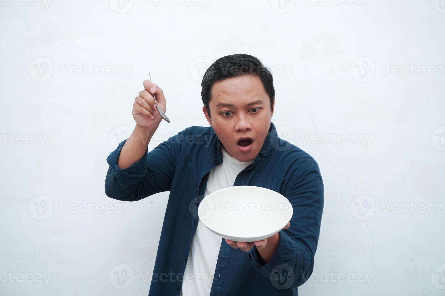 Portrait of happy Asian man wearing a blue shirt while holding an empty plate with happy and excited expression. Young man showing and pointing empty mockup plate. Isolated with a white background. photo