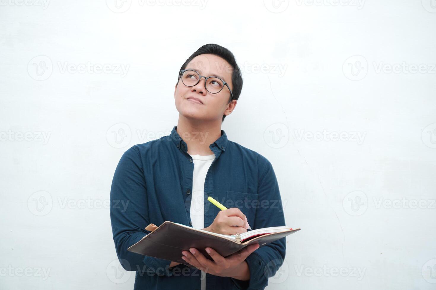 Young Asian man holding a book while thinking and concentrating. Asian student looking for ideas while studying over white background photo