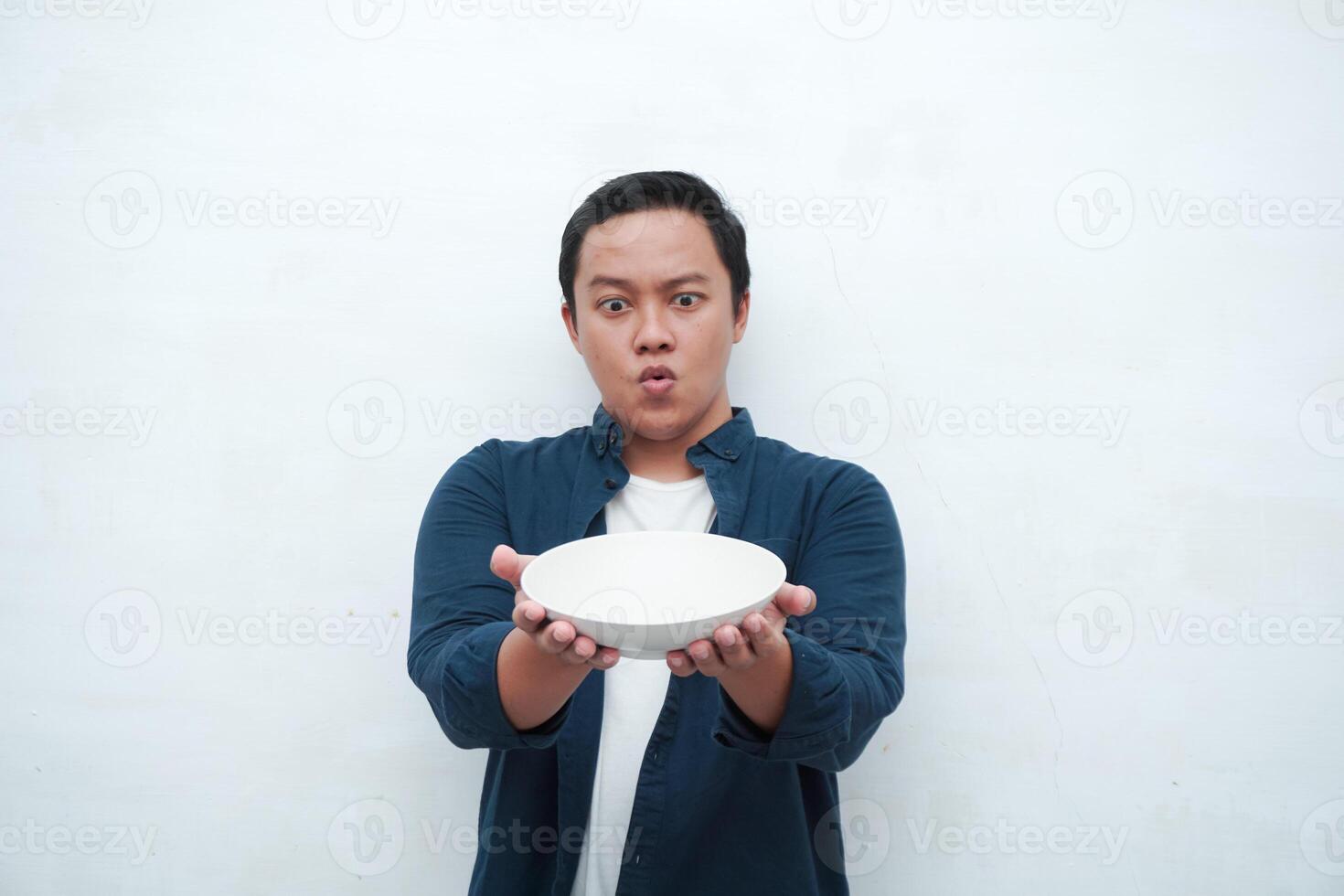 Portrait of happy Asian man wearing a blue shirt while holding an empty plate with happy and excited expression. Young man showing and pointing empty mockup plate. Isolated with a white background. photo