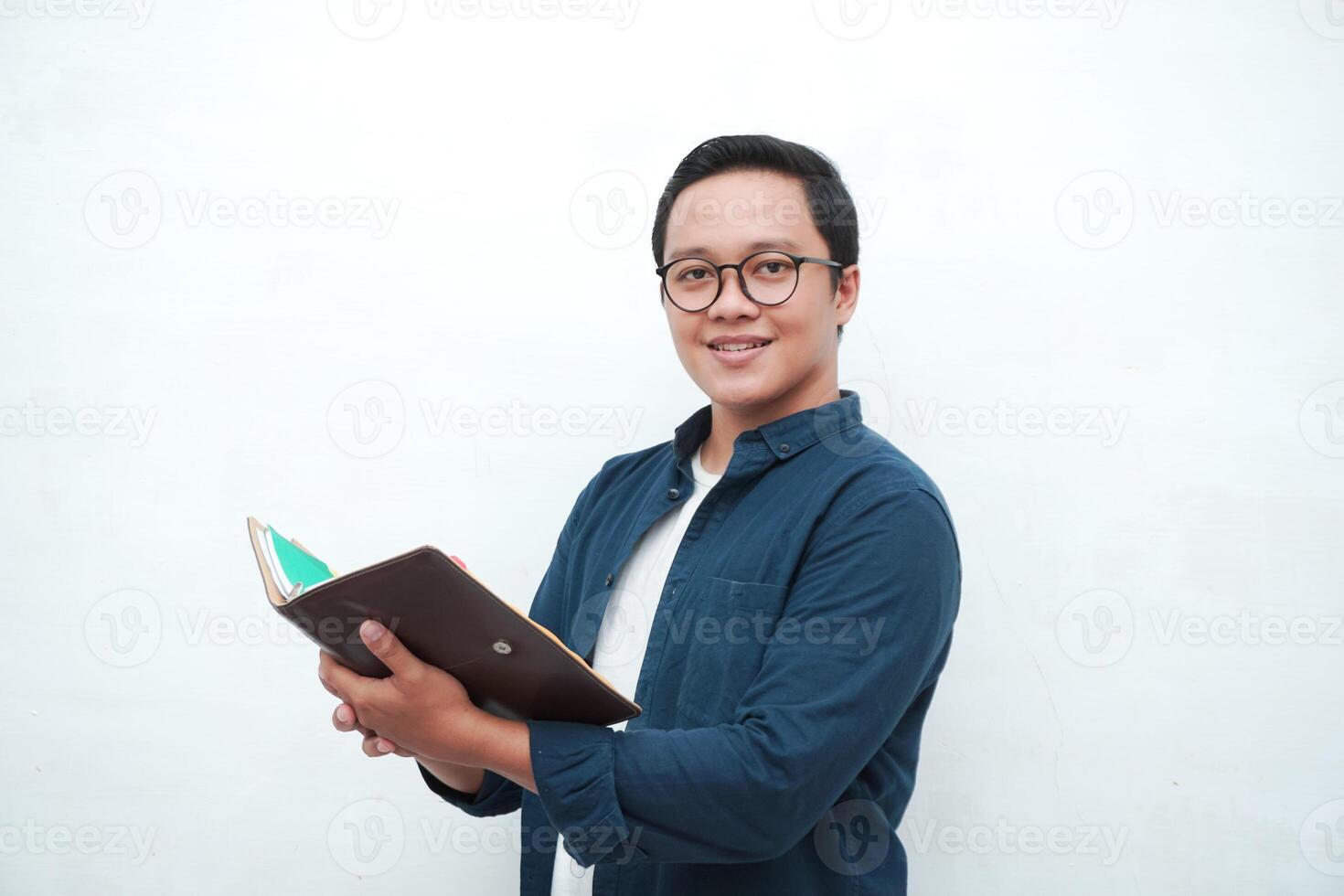 Young Asian man holding books smiling happy and positive while reading a book. Asian student flipping a book page over white background photo