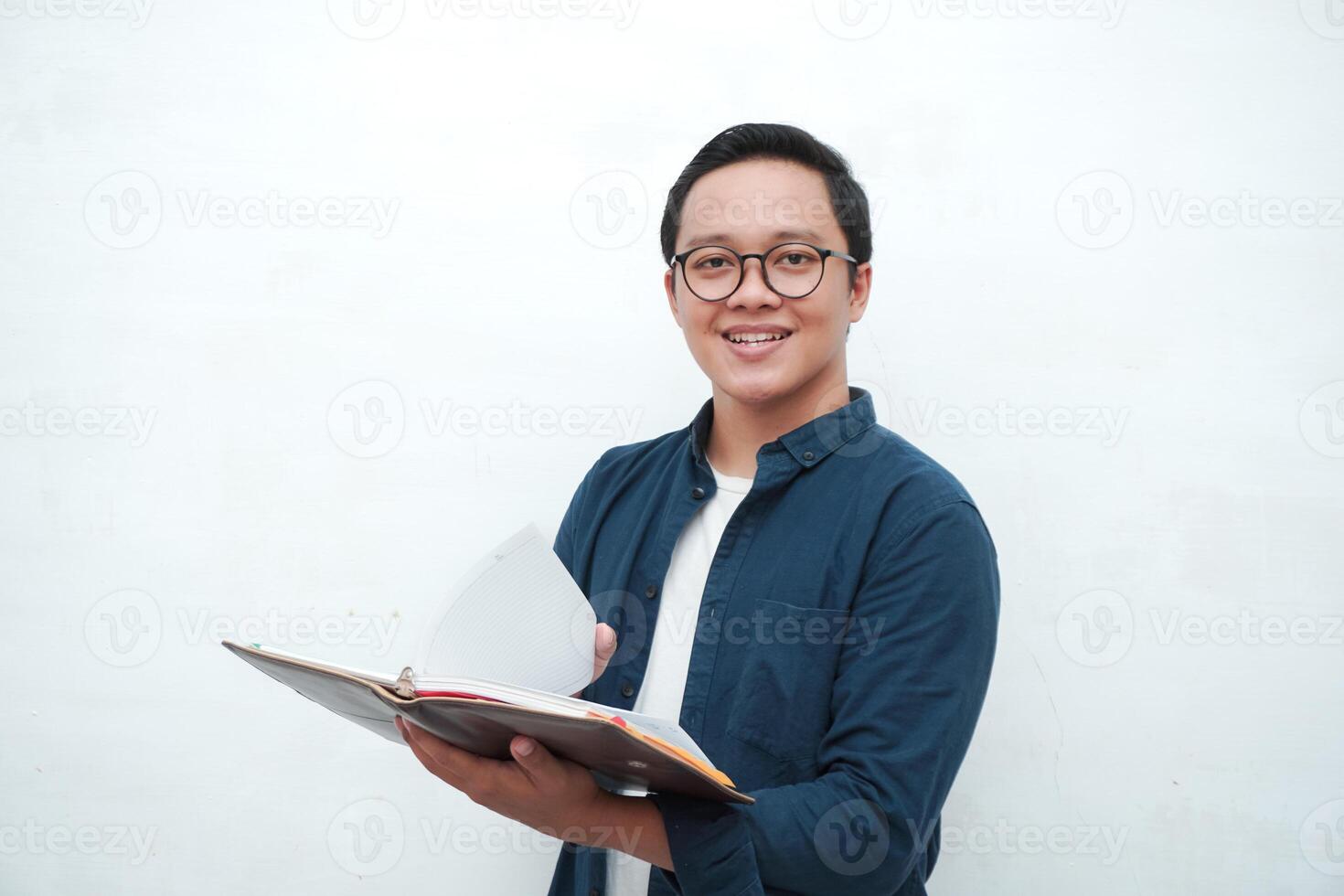 Young Asian man holding books smiling happy and positive while reading a book. Asian student flipping a book page over white background photo