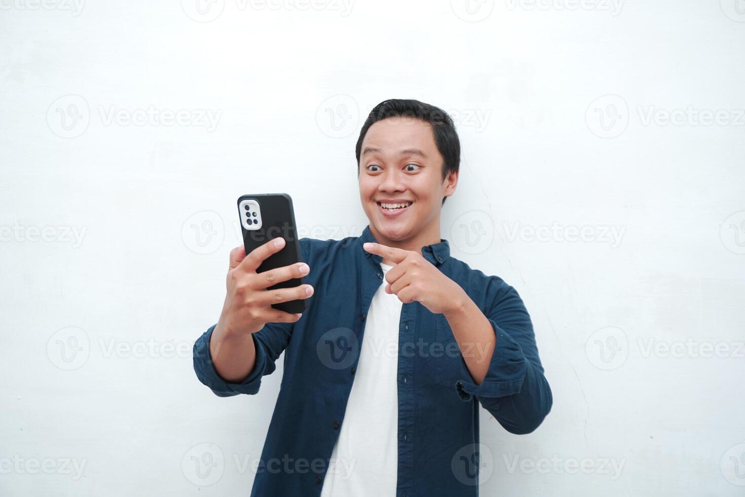 Young Asian man looking smartphone with shocked and surprised expression. Male with blue t-shirt showing excited expression isolated on white background photo