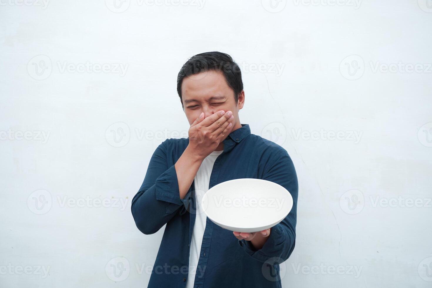 Asian man covering his mouth and nose while holding an empty mockup plate. Covering mouth and nose because of bad smell of food photo