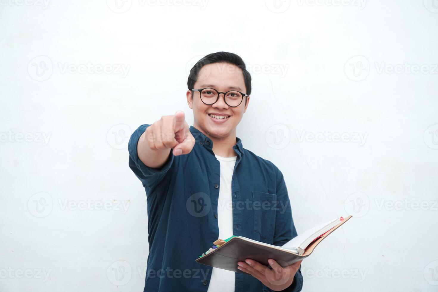 Young Asian man holding books smiling happy and positive while reading a book. Asian student pointing and showing welcoming gesture photo