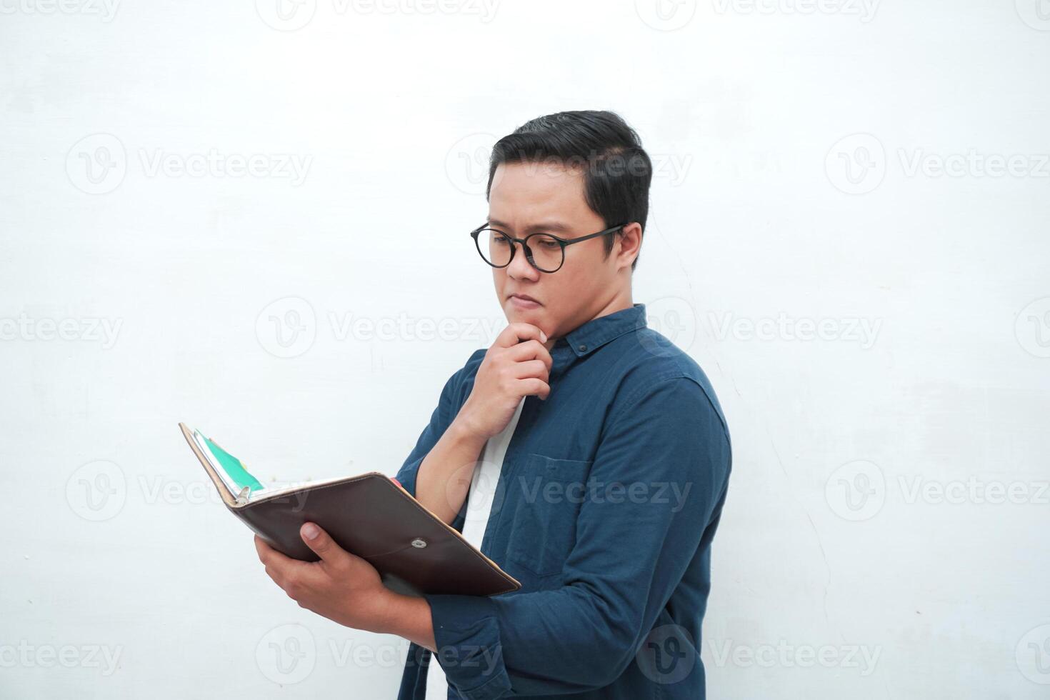 Young Asian man holding a book while thinking and concentrating. Asian student looking for ideas while studying over white background photo