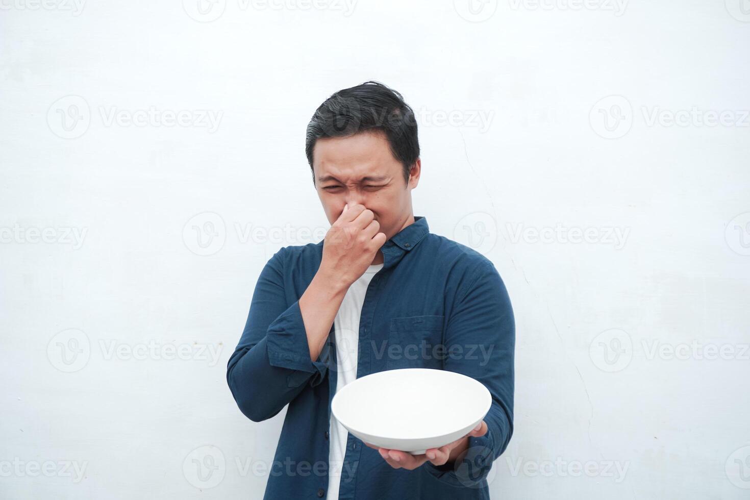 Asian man covering his mouth and nose while holding an empty mockup plate. Covering mouth and nose because of bad smell of food photo