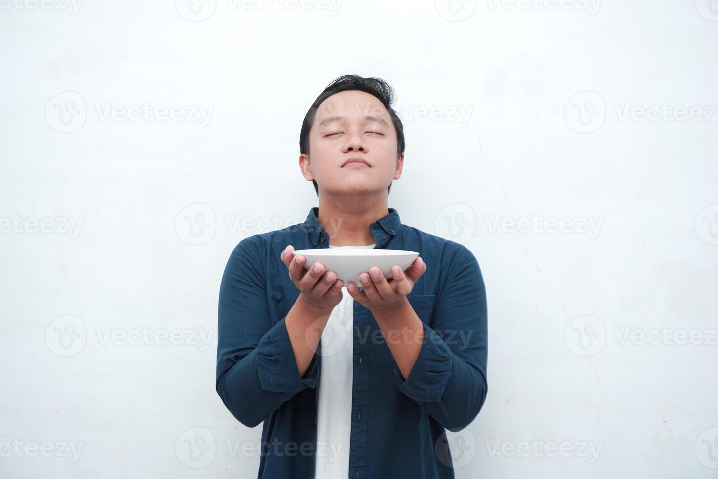A portrait of happy Asian man wearing a blue shirt excited to eat a plate of fried noodles. Young man smelling the aroma of delicious fried noodles. Isolated with a white background. photo