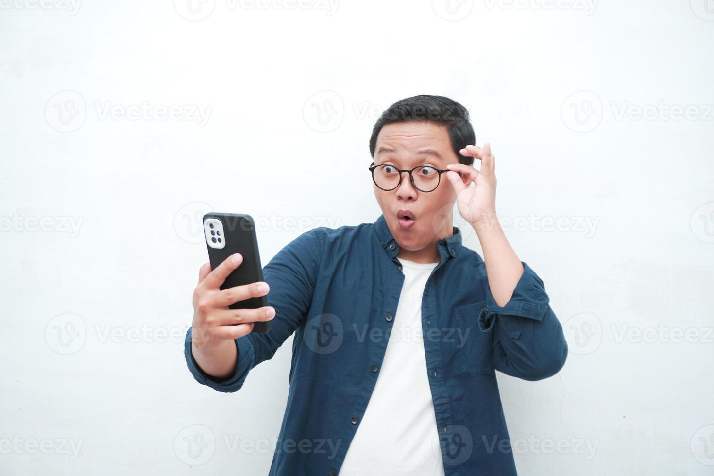 Young Asian man looking smartphone with shocked and surprised expression. Male with blue t-shirt showing excited expression isolated on white background photo