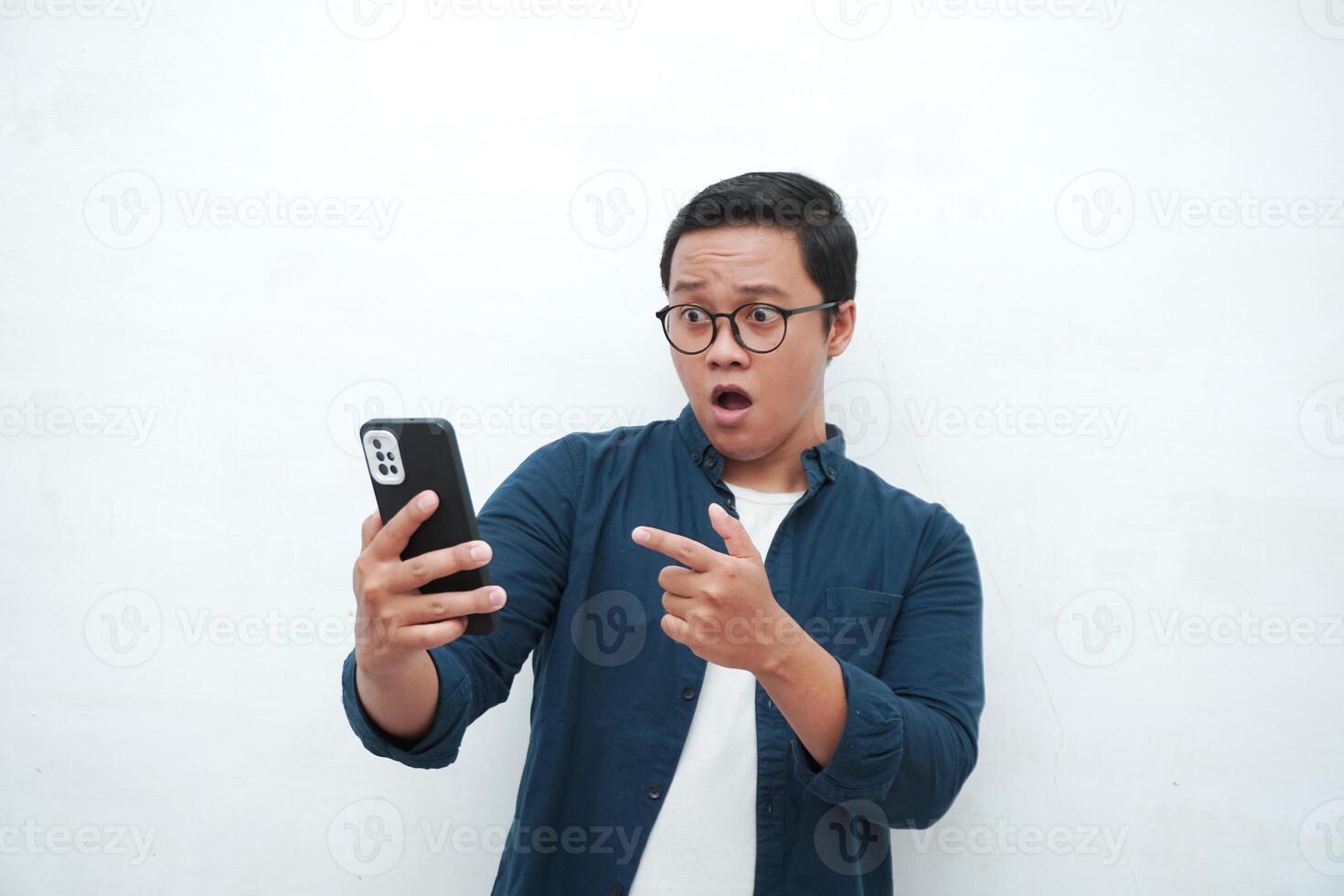 Young Asian man looking smartphone with shocked and surprised expression. Male with blue t-shirt showing excited expression isolated on white background photo