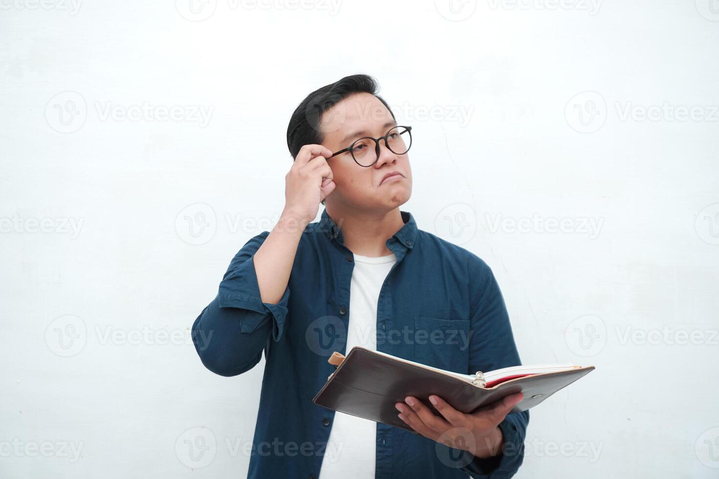 Young Asian man holding a book while thinking and concentrating. Asian student looking for ideas while studying over white background photo