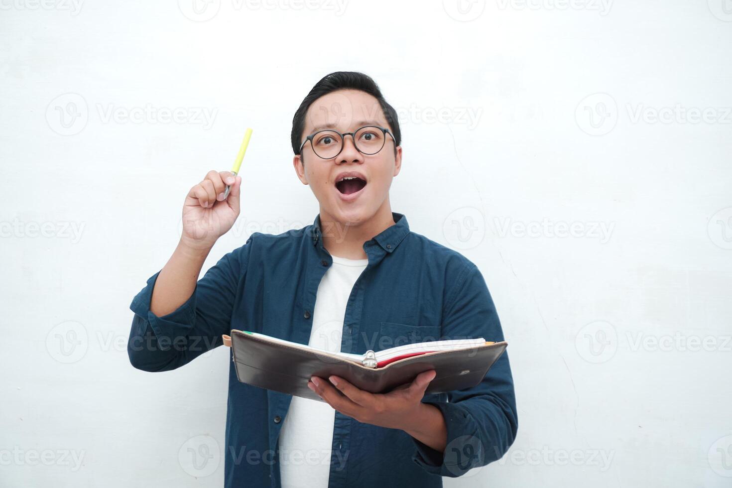 Happy young Asian man with glasses showing expression of gets genius idea. Asian college student showing happy and excited expression while holding book and pen photo
