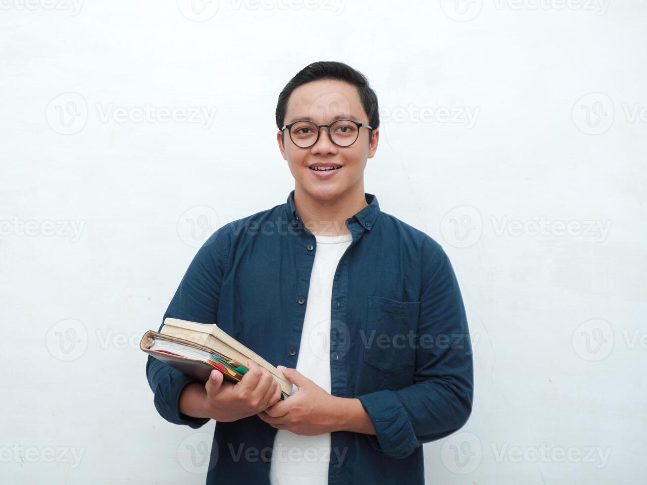 Young Asian man holding books smiling happy and positive while reading a book. Asian student flipping a book page over white background photo