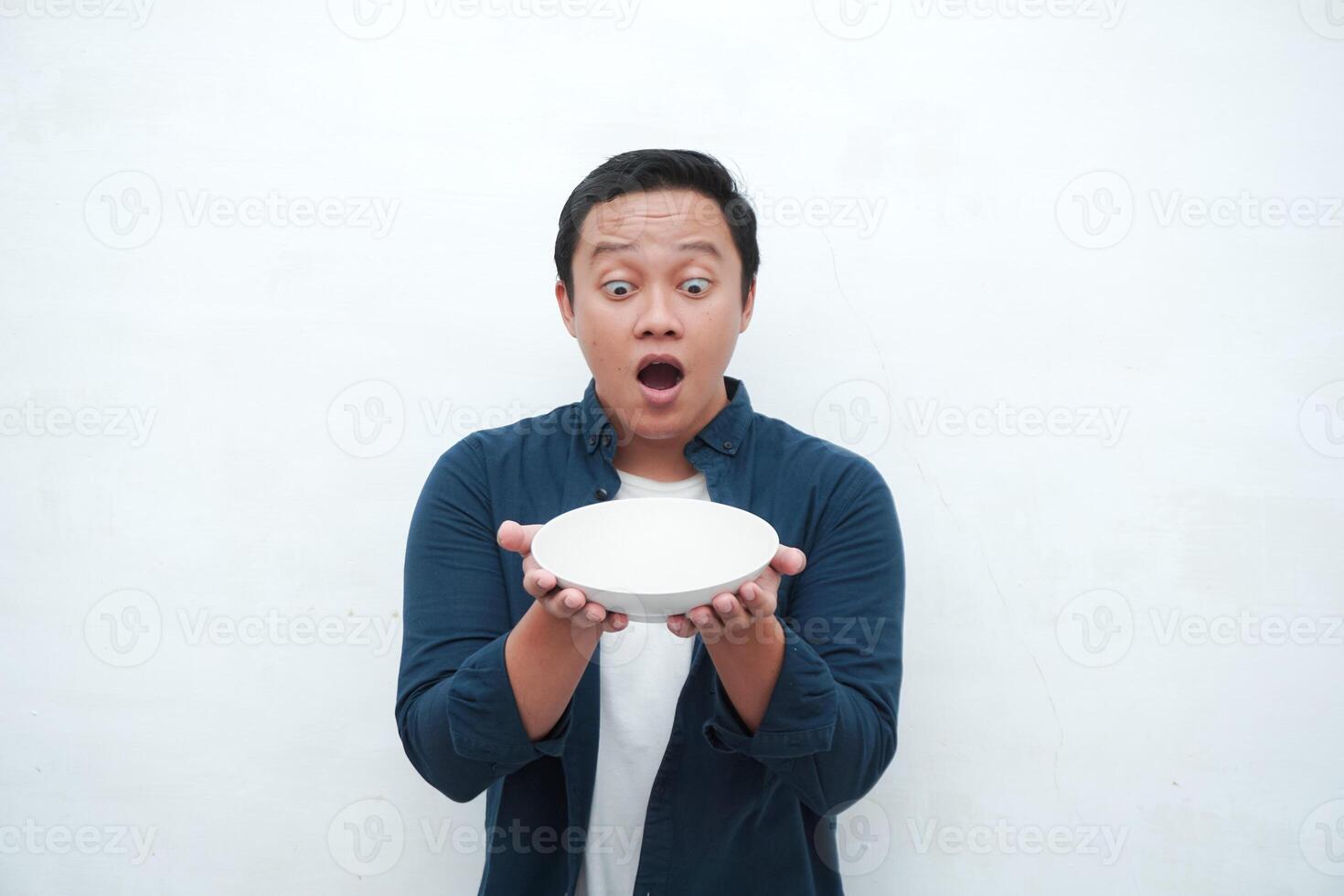 Portrait of happy Asian man wearing a blue shirt while holding an empty plate with happy and excited expression. Young man showing and pointing empty mockup plate. Isolated with a white background. photo