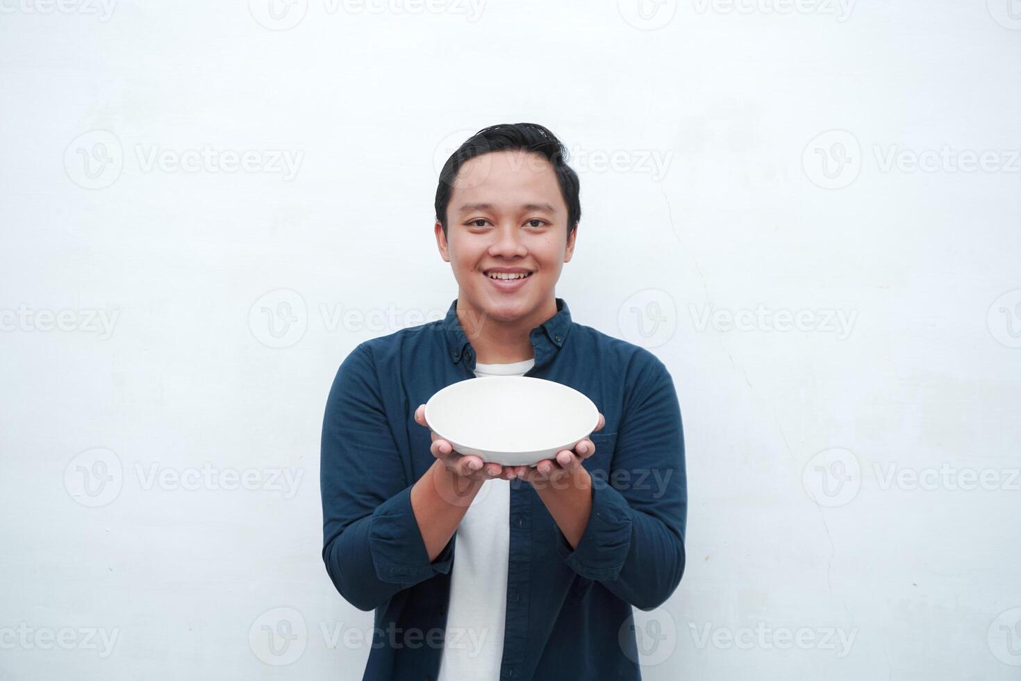 Portrait of happy Asian man wearing a blue shirt while holding an empty plate with happy and excited expression. Young man showing and pointing empty mockup plate. Isolated with a white background. photo