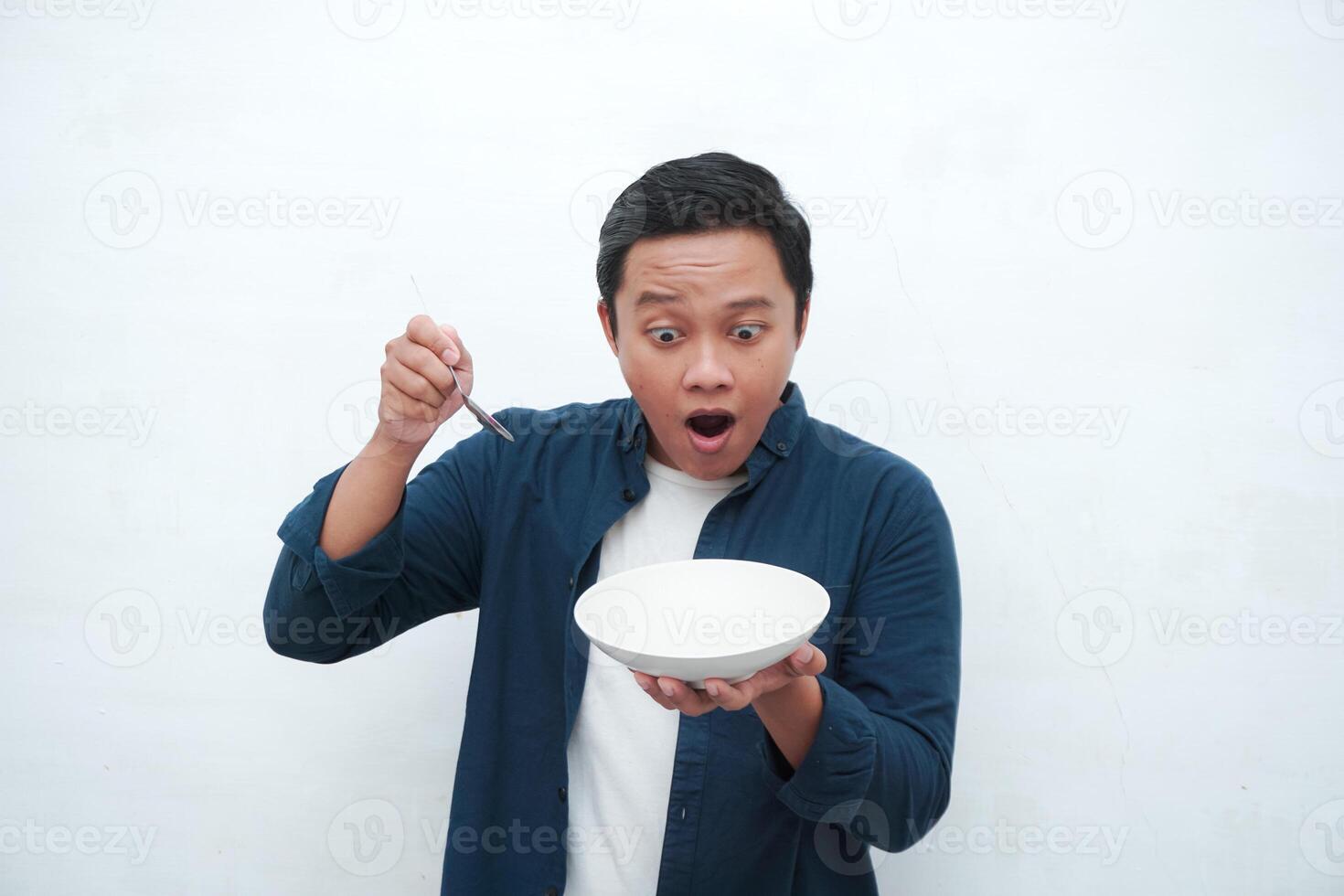 Portrait of happy Asian man wearing a blue shirt while holding an empty plate with happy and excited expression. Young man showing and pointing empty mockup plate. Isolated with a white background. photo