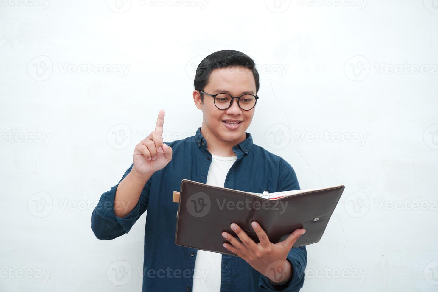 Happy young Asian man with glasses showing expression of gets genius idea. Asian college student showing happy and excited expression while holding book and pen photo