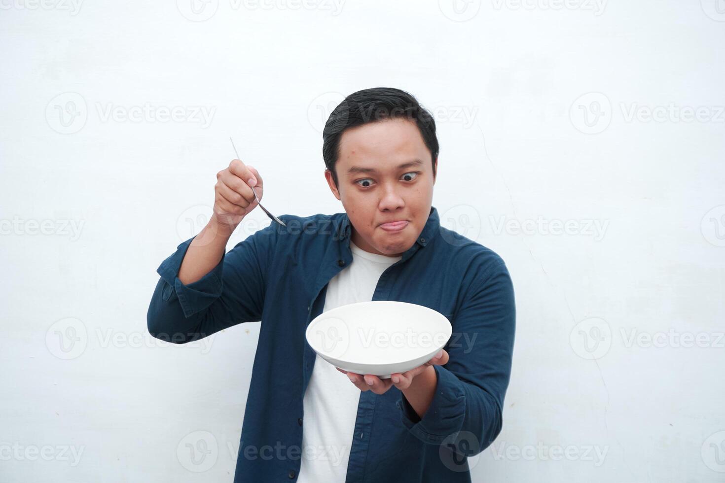 Portrait of happy Asian man wearing a blue shirt while holding an empty plate with happy and excited expression. Young man showing and pointing empty mockup plate. Isolated with a white background. photo