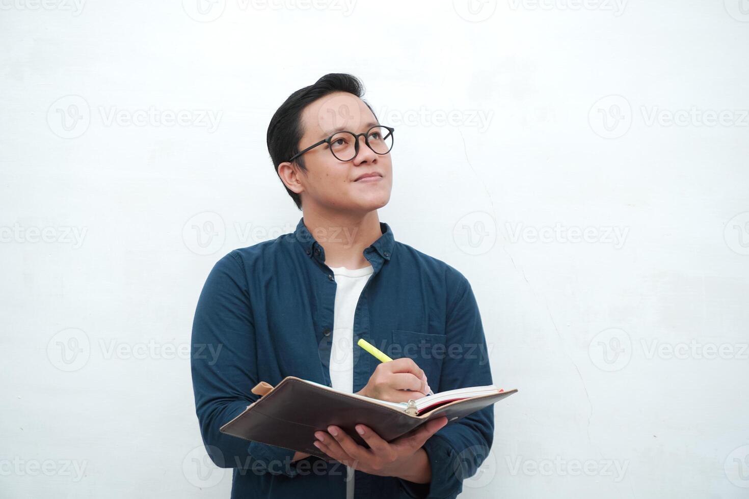 Happy young Asian man with glasses showing expression of gets genius idea. Asian college student showing happy and excited expression while holding book and pen photo