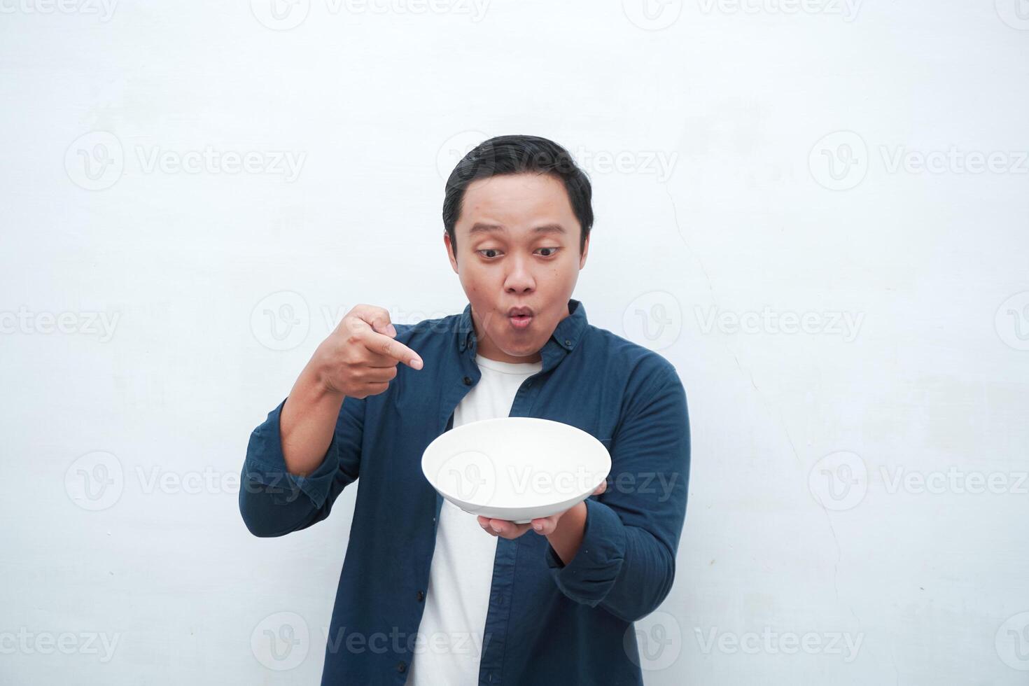 Portrait of happy Asian man wearing a blue shirt while holding an empty plate with happy and excited expression. Young man showing and pointing empty mockup plate. Isolated with a white background. photo