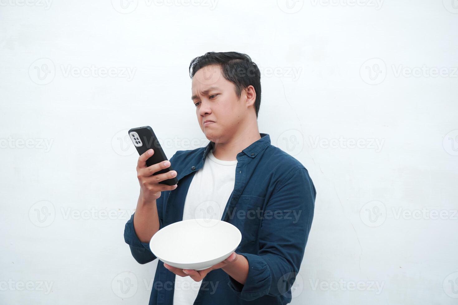 A portrait of happy Asian man wearing a blue shirt while thinking what to eat. Young man holding spoon and empty mockup plate. Isolated with a white background. photo
