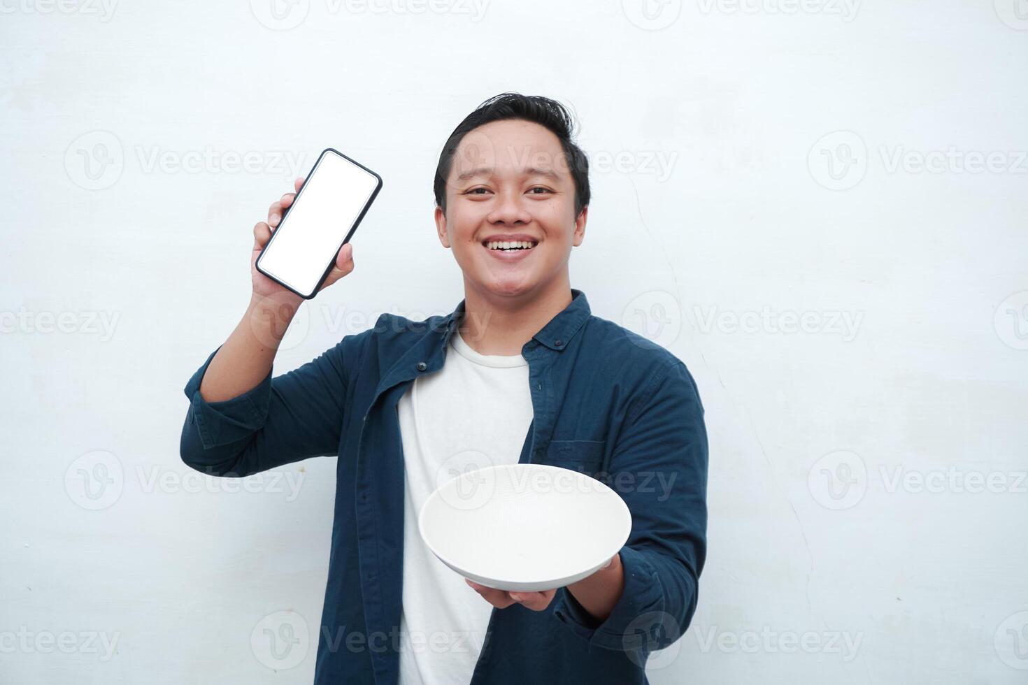 Happy Asian man holding blank smartphone screen and white plate with happy and excited expression. Asian man holding empty white plate smiling and showing white mobile phone screen for advertisement. photo