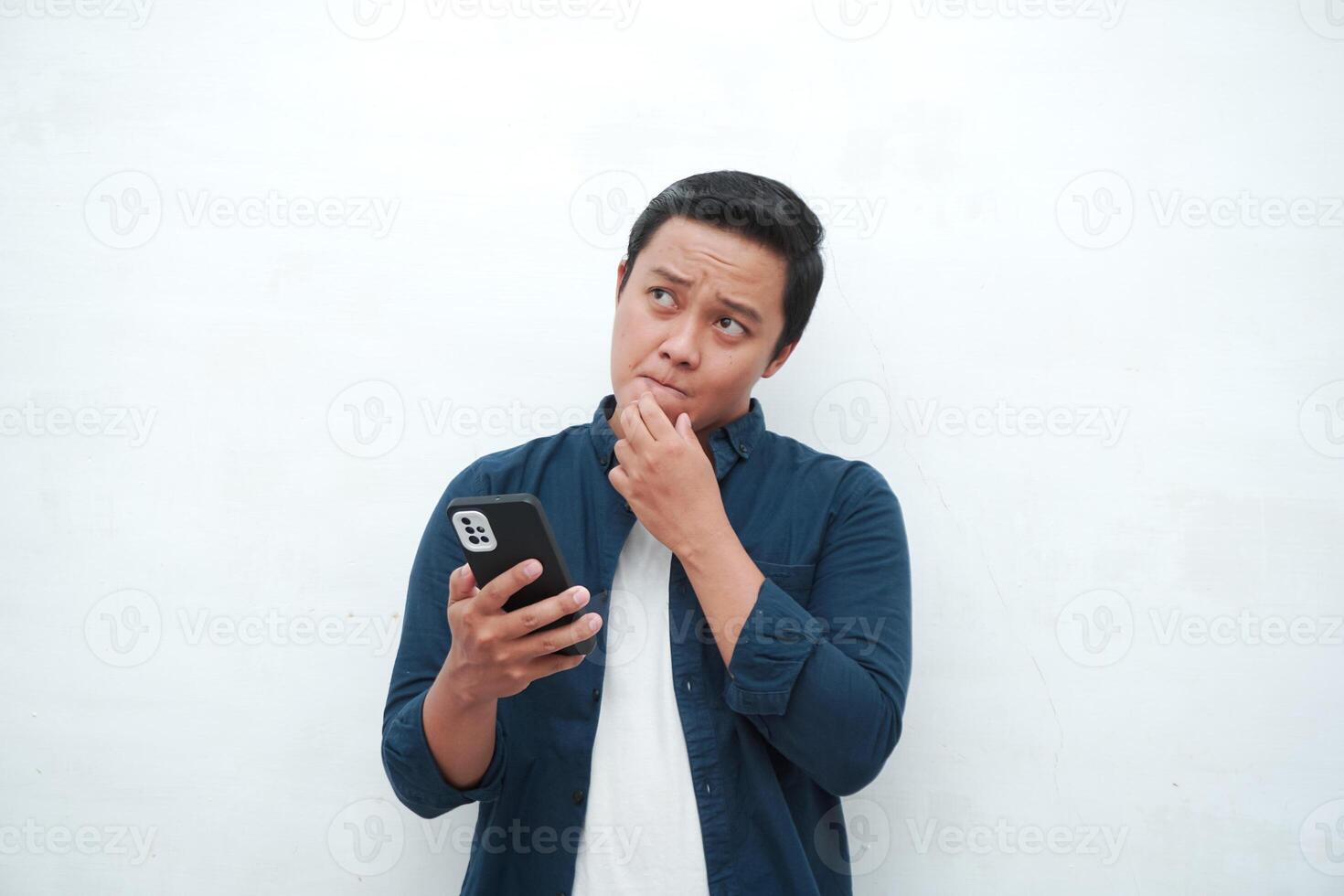 Young Asian man thinking and concentrating while holding books and handphone. Asian student thinking and looking for ideas while holding phone over white background photo