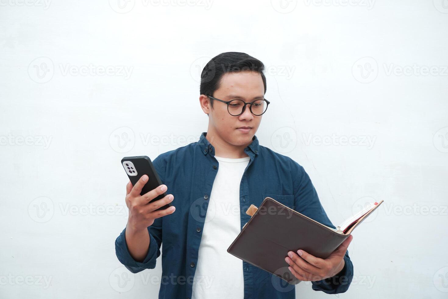 Asian college student holding book and blank smartphone screen with happy and excited expression. Young Asian student wearing blue shirt smiling and showing white mobile phone screen photo