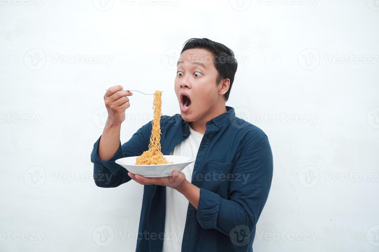 A portrait of happy Asian man wearing a blue shirt while holding a plate of fried noodles. Young man smiling and pointing to a plate of noodle. Isolated with a white background. photo