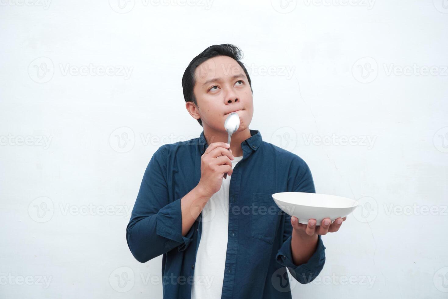 A portrait of happy Asian man wearing a blue shirt while thinking what to eat. Young man holding spoon and empty mockup plate. Isolated with a white background. photo
