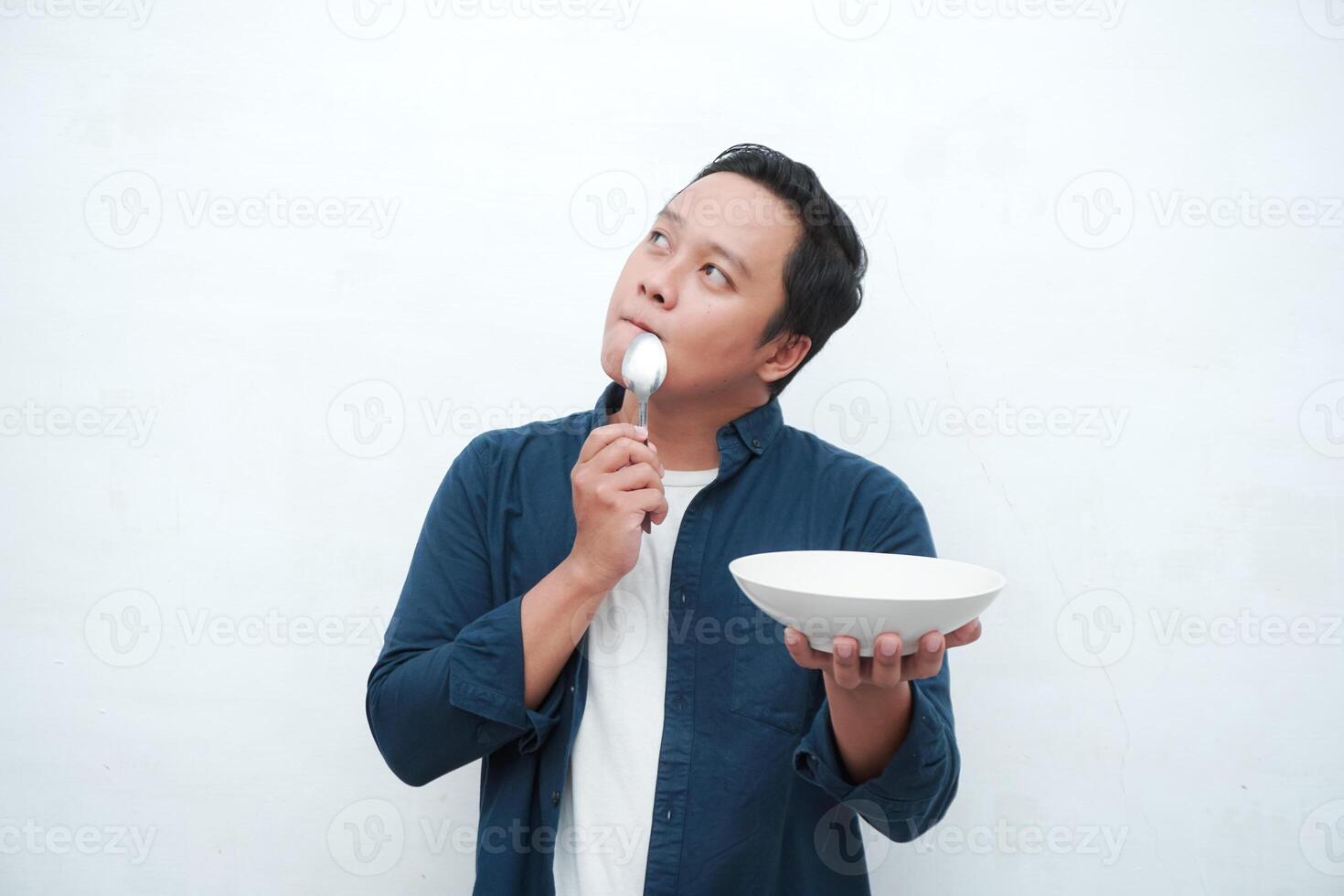 A portrait of happy Asian man wearing a blue shirt while thinking what to eat. Young man holding spoon and empty mockup plate. Isolated with a white background. photo