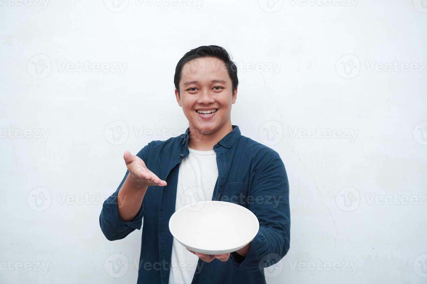Portrait of happy Asian man wearing a blue shirt while holding an empty plate with happy and excited expression. Young man showing and pointing empty mockup plate. Isolated with a white background. photo