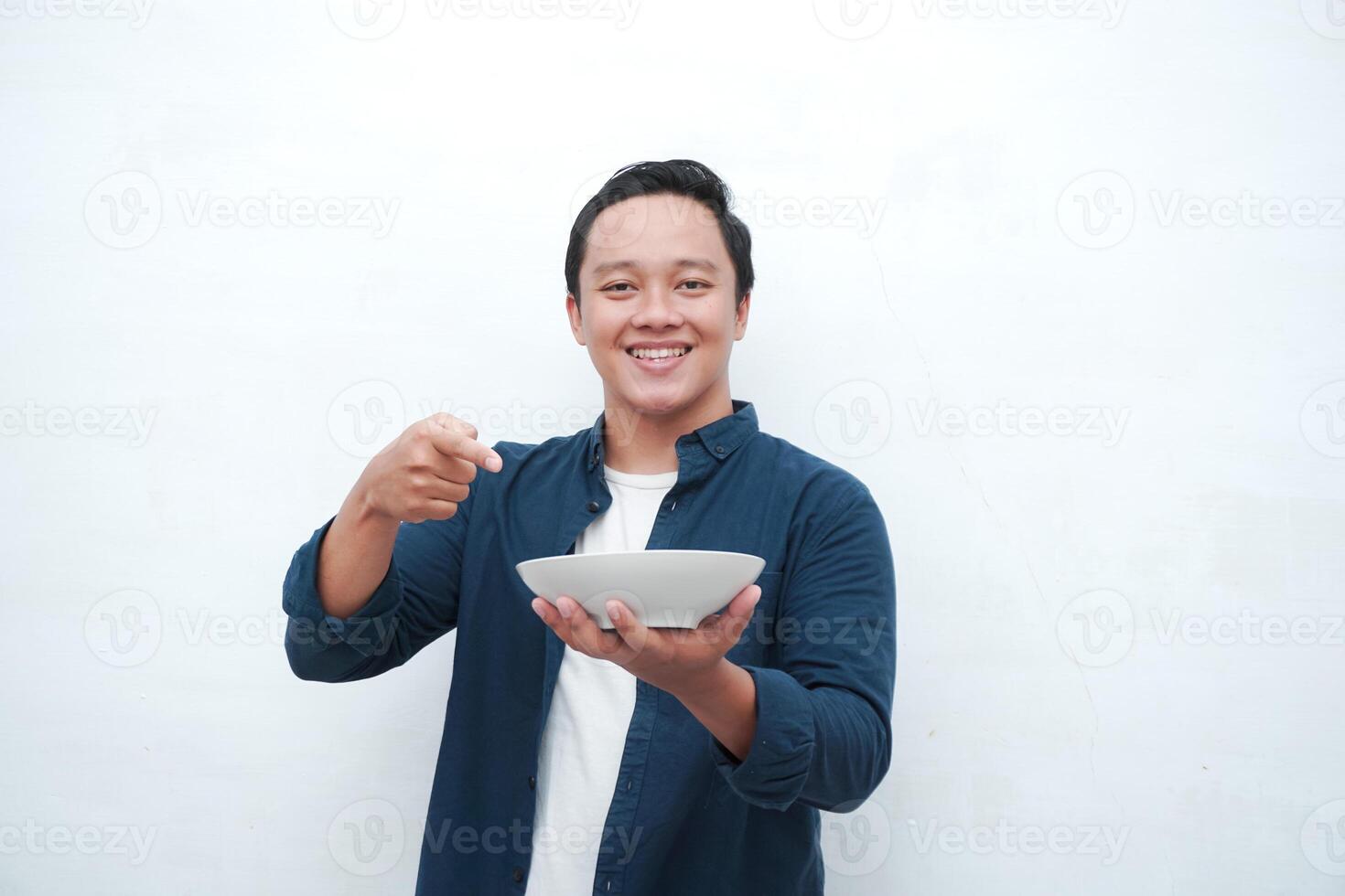 Portrait of happy Asian man wearing a blue shirt while holding an empty plate with happy and excited expression. Young man showing and pointing empty mockup plate. Isolated with a white background. photo