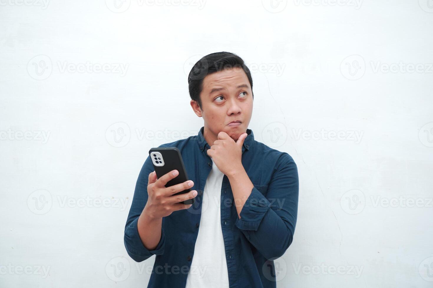 Young Asian man thinking and concentrating while holding books and handphone. Asian student thinking and looking for ideas while holding phone over white background photo