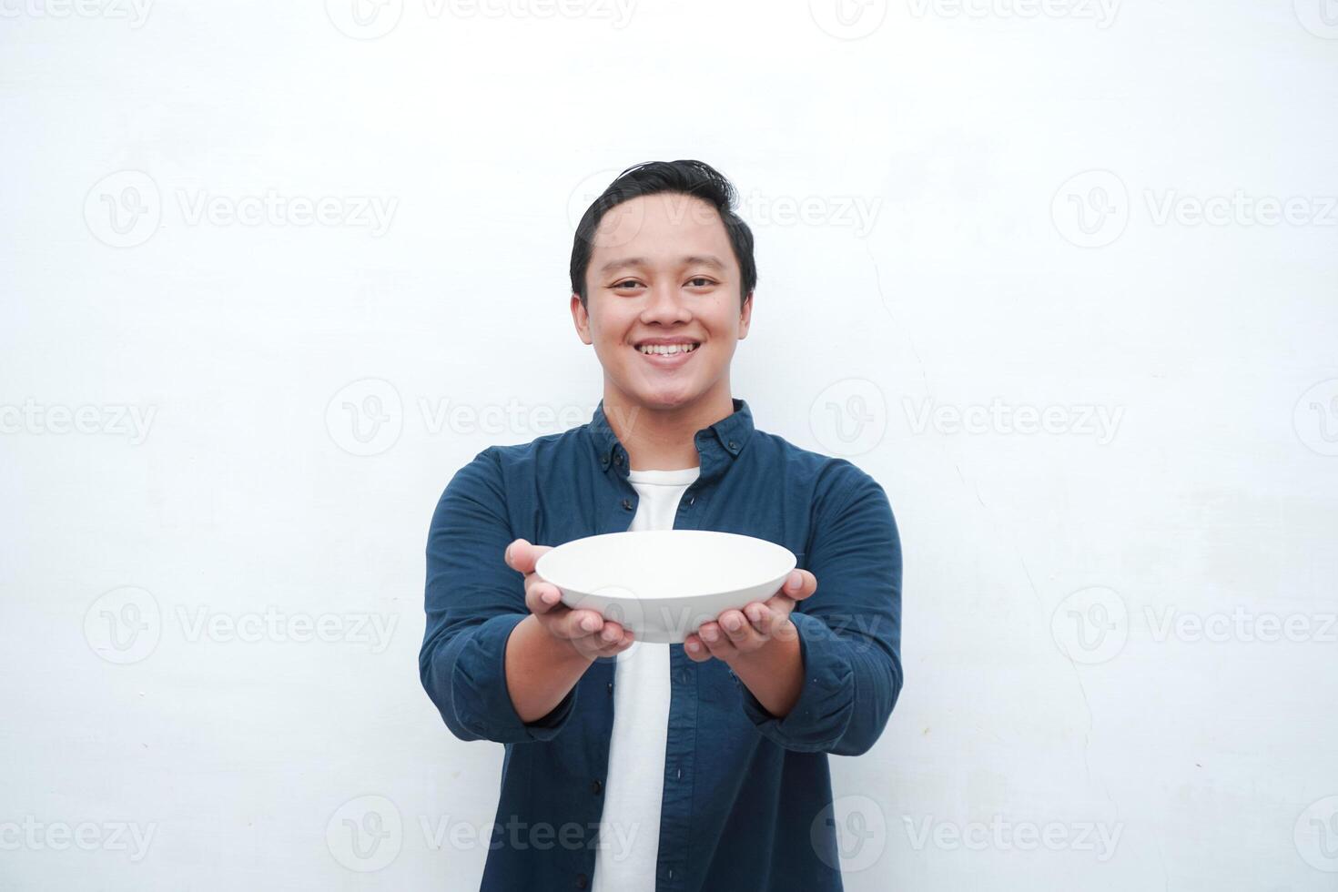 Portrait of happy Asian man wearing a blue shirt while holding an empty plate with happy and excited expression. Young man showing and pointing empty mockup plate. Isolated with a white background. photo