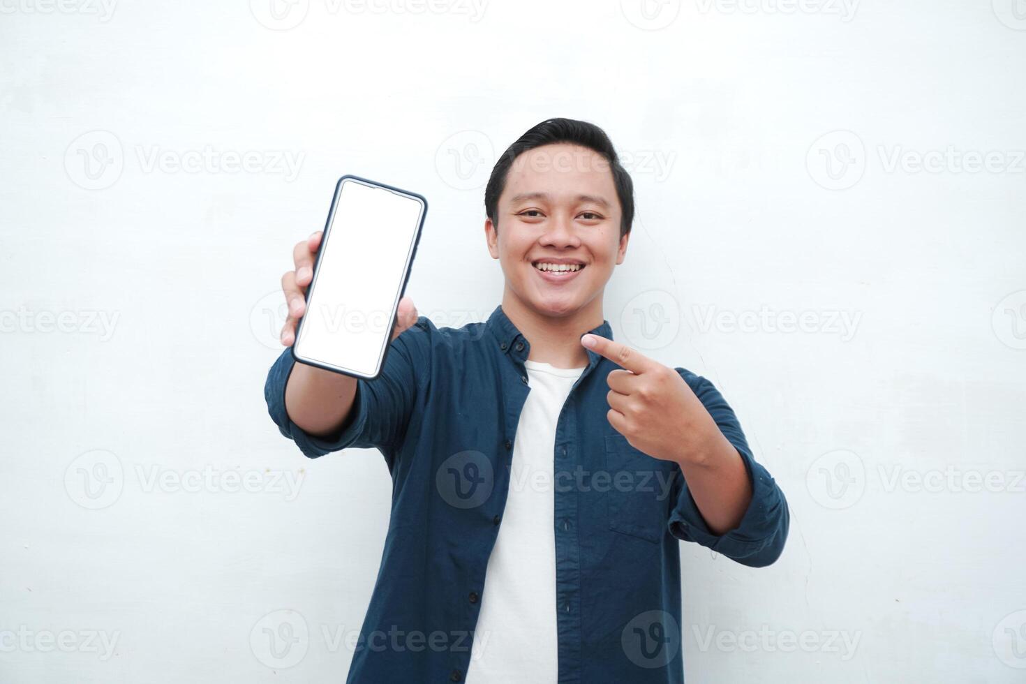 Happy Asian man holding blank smartphone screen with happy and excited expression. Young Asian man wearing blue shirt smiling and showing white mobile phone screen for advertisement. photo