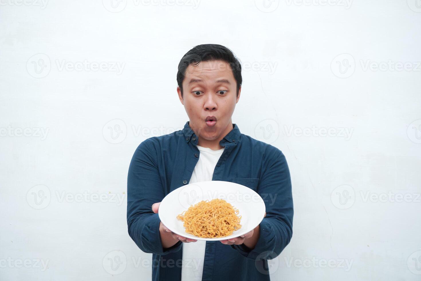 A portrait of happy Asian man wearing a blue shirt while holding a plate of fried noodles. Young man smiling and pointing to a plate of noodle. Isolated with a white background. photo