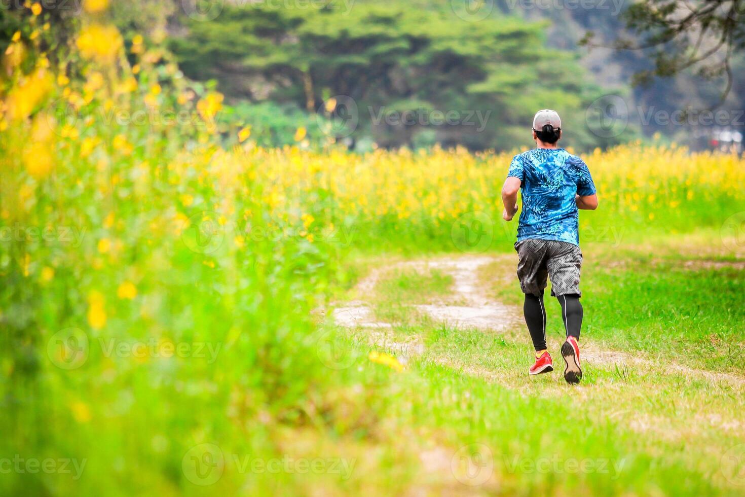 Man Running on a Trail in a Lush Green Field with Yellow Flowers and Trees in the Background photo