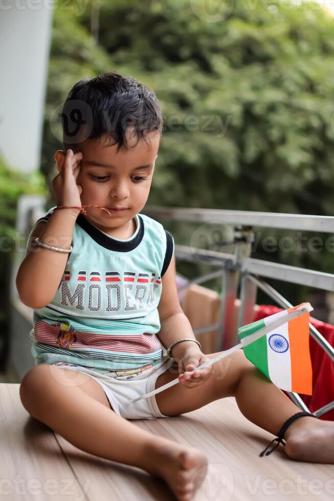 Cute Indian boy holding Indian National Flag in his hangs at home balcony. Bright portrait of happy child sitting on the table with national flag photo